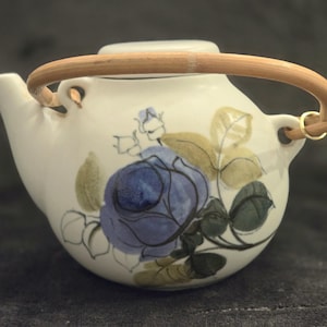 May include: White ceramic teapot with a bamboo handle and a lid. The teapot is decorated with a blue rose and green and tan leaves. The handle is attached with gold-colored metal rings.