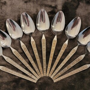 May include: A set of 10 silver spoons with ornate gold detailing on the handles. The spoons are arranged in a fan shape, with the handles pointing outwards.