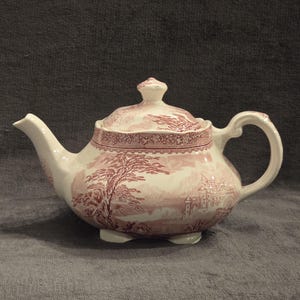 May include: A white ceramic teapot with a lid, featuring a red and white landscape design with trees and a castle. The teapot has a curved spout, a rounded body, and a loop handle. The teapot sits on three small feet.