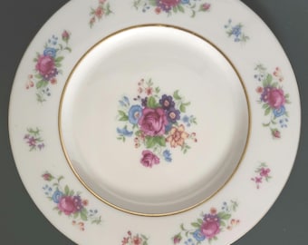 May include: A round white porcelain plate featuring a central floral bouquet of pink, blue, and orange flowers with green leaves. The rim is decorated with smaller matching floral clusters and finished with a thin gold band around the inner edge.