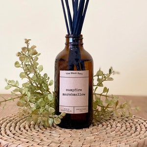 May include: A brown glass reed diffuser bottle with black reeds. The label reads "Campfire Marshmallow" from The Black Barn. The diffuser is surrounded by greenery and sits on a woven mat.