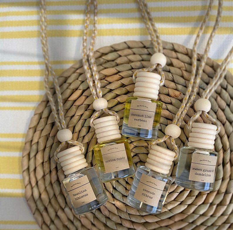 May include: An overhead shot of five small glass bottles with wooden caps and hanging cords. Each bottle contains a different colored liquid and has a label with a scent name: Hawaiian Hibiscus, Cotton Candy, Avocado Citrus, Sweet Grass & Dandelion, and Coconut Lime Verbena.