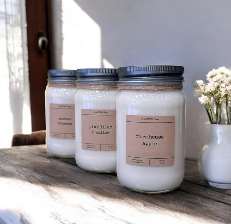 May include: Three glass mason jar candles with white wax and dark metal lids. Each jar features a tan rectangular label with black text reading The Black Barn and scent names: cactus blossom, pink lilac & willow, and farmhouse apple.