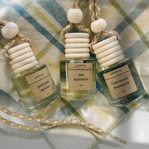 May include: Three small glass bottles with wooden tops and hanging cords. Each bottle has a label with a scent name: "poolside splash", "sea minerals", and "lime basil mandarin". The bottles are filled with colored liquid and rest on a checkered cloth.