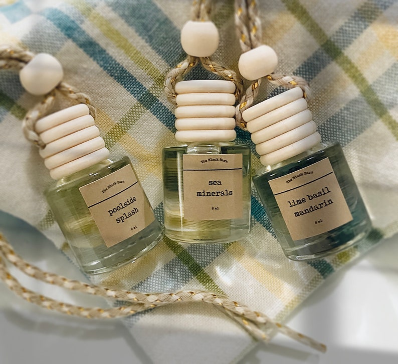 May include: Three small glass bottles with wooden tops and hanging cords. Each bottle has a label with a scent name: "poolside splash", "sea minerals", and "lime basil mandarin". The bottles are filled with colored liquid and rest on a checkered cloth.