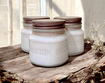 May include: Three white mason jar candles with brown lids and twine accents. The jars are filled with white wax and sit on a rustic wooden surface. The word "MASON" is embossed on the front of the jars.