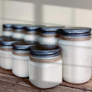 May include: A collection of clear glass jars filled with white wax, each topped with a dark metal lid. A piece of brown twine is wrapped around the neck of each jar. The jars are arranged in a group, with some appearing larger than others.