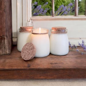 May include: Three glass jar candles with cork lids. One candle is lit, and the flame is visible. The jars are filled with white wax and sit on a wooden surface. A cork lid rests in front of the candles.
