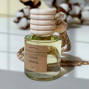 May include: A clear glass bottle with a wooden cap and a label that reads "clean cotton" and "8 ml." The bottle is filled with a light yellow liquid and has a decorative rope and bead attached. The brand name "The Black Barn" is also visible.