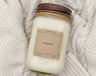 Puede incluir: Una vela de cera de soja blanca en un tarro con tapa marrón. La etiqueta dice "winter" y "The Black Barn". El tarro está envuelto con hilo y descansa sobre una manta de punto color crema. La vela es de 473 ml.