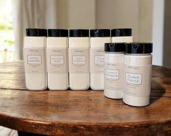 May include: A collection of white carpet and upholstery freshener containers with black lids. Each container has a label with a different scent name: Lemon Lavender, Farmhouse Apple, Sweet Strawberry, Clear Cotton, Summer Pineapple, and Guava Fresca.