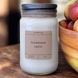 May include: A clear glass mason jar candle with a black metal lid and a twine wrap around the neck. The label reads The Black Barn, farmhouse apple, hand poured, nashville, in, soy wax, 16 oz. The candle contains white wax and is filled to the top.