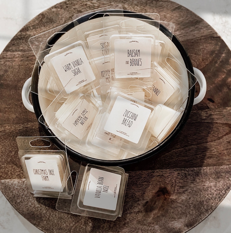 May include: Scented wax melts in clear plastic packaging, presented in a white and black enamel bowl. Labels include text like "Warm Vanilla Sugar," "Balsam and Berries," and "Zucchini Bread." The bowl sits on a wooden surface.