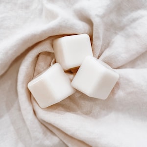 May include: Three white, square objects with rounded edges are arranged on a cream-coloured, textured fabric. The objects appear smooth and glossy, reflecting the light. The fabric is draped, creating soft folds and shadows.