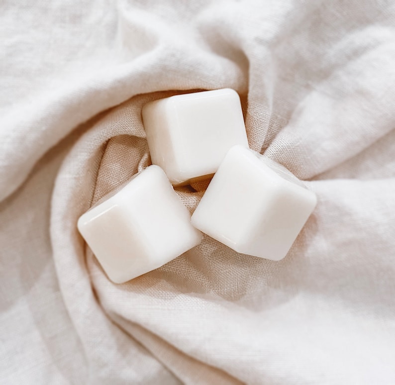 May include: Three white, square objects with rounded edges are arranged on a cream-coloured, textured fabric. The objects appear smooth and glossy, reflecting the light. The fabric is draped, creating soft folds and shadows.
