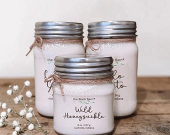 May include: Three glass mason jars filled with light pink wax, each topped with a silver metal lid and wrapped with brown twine. The front label reads Wild Honeysuckle, 8 oz | 227 g, the Black Barn, and Nashville, Indiana in black text.