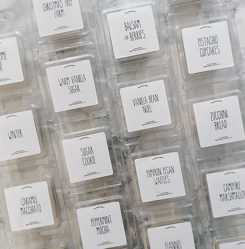 May include: Overhead view of wax melt cubes in clear plastic containers. Each cube has a white label with black text, featuring scents such as "Balsam and Berries" and "Zucchini Bread". The labels are neatly arranged.