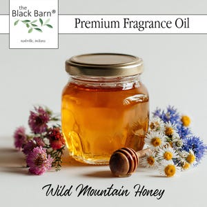 May include: A small glass jar filled with golden-colored Wild Mountain Honey, topped with a gold lid. A wooden honey dipper rests beside the jar, surrounded by pink, white, and blue flowers. The text "Premium Fragrance Oil" and "Wild Mountain Honey" are displayed.