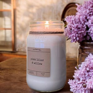 May include: A clear glass mason jar candle with a white wax fill and a brown paper label reading The Black Barn, pink lilac & willow, hand poured, Nashville, IN, soy wax, 16 oz. The jar neck is wrapped in twine and the candle is lit.