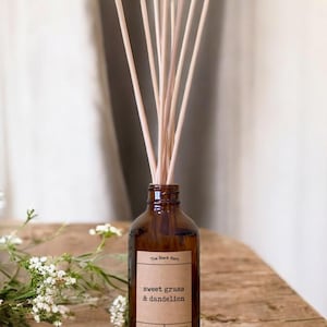 May include: A brown glass diffuser bottle with wooden reeds. The label reads "sweet grass & dandelion" from "The Black Barn". The bottle sits on a wooden surface with white flowers in the background. The diffuser is a home fragrance product.