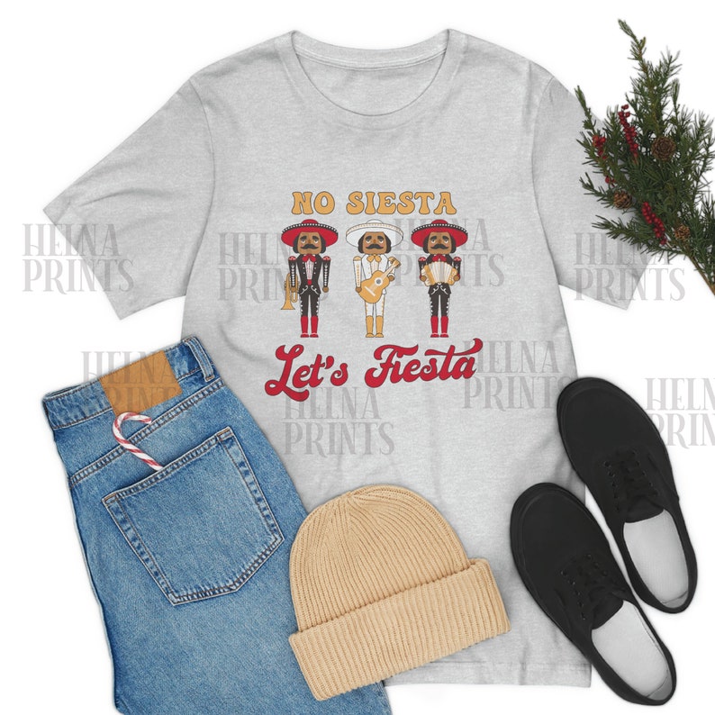 May include: A gray t-shirt with the text "NO SIESTA Let's Fiesta" in red and white. Three cartoon mariachi nutcrackers are depicted above the text. The t-shirt is displayed on a flat surface with a pair of black shoes, a beanie, and blue jeans.