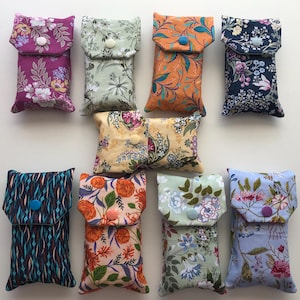 May include: A collection of nine rectangular fabric pouches featuring various floral and abstract patterns in colours including purple, sage green, orange, navy blue, yellow, and light blue, each secured with a single plastic snap button closure.