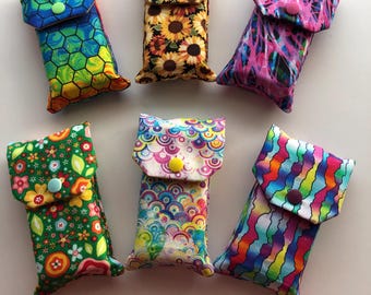 May include: Six rectangular fabric pouches with fold-over flaps secured by plastic snap buttons. The pouches feature various colourful patterns including sunflowers, geometric honeycombs, abstract paint splatters, floral prints, and rainbow waves.