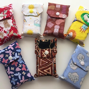 May include: Seven rectangular fabric pouches with snap closures, featuring various patterns including red floral, yellow suns, brown yarn balls, yellow animal prints, blue butterflies, black and brown geometric shapes, and light blue elephant motifs.