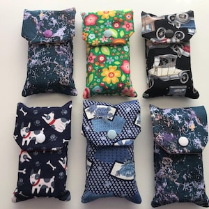 May include: Six rectangular fabric pouches with snap closures, featuring various patterns including a dark marble print, a bright floral design, vintage cars on black, cartoon dogs, blue geometric fan motifs, and a dark abstract textured pattern.