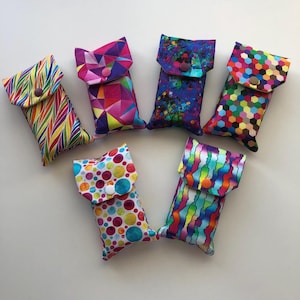 May include: Six rectangular fabric pouches with snap closures, each featuring a distinct colourful pattern including diagonal stripes, geometric shapes, paint splatters, multicoloured hexagons, polka dots, and wavy vertical lines in vibrant hues.