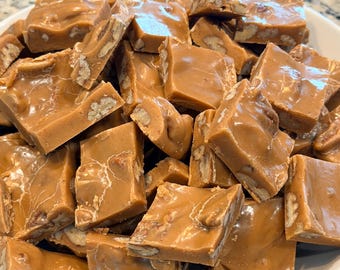 Peut inclure: Un bol blanc rempli de nombreux morceaux carrés de fudge au caramel brun clair. Chaque morceau de fudge lisse et brillant contient des morceaux de noix de pécan visibles incrustés dans la confiserie, créant un aspect texturé dans le tas.