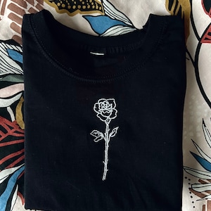 May include: Black t-shirt with a white embroidered rose design.