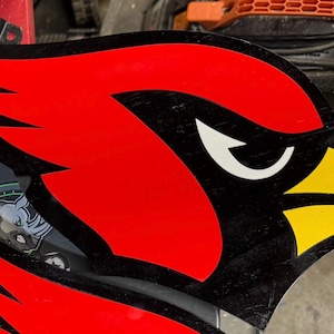 Puede incluir: Recorte de madera de la cabeza de un pájaro cardenal, con plumas rojas vibrantes, un contorno negro y un pico amarillo. El ojo del pájaro es blanco. El diseño es probablemente para decoración deportiva o de equipo.