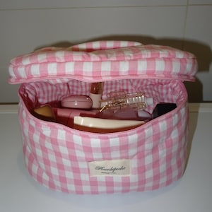 May include: A pink and white gingham cosmetic bag, open to show various beauty products. The bag has a handle and a zip closure. The label reads "Fleurdepoche Handmade in France."