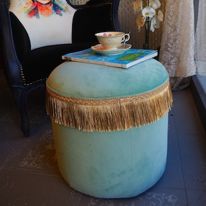 Puede incluir: Un otomano de terciopelo azul verdoso con ribete de flecos dorados y un libro titulado "Anne Stokes Paints Everything" en la parte superior. Una taza de té blanca y un platillo con flores rosas están en un plato azul y blanco al lado del libro.