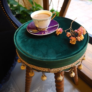 Könnte beinhalten: Ein grüner Samt-Hocker mit goldenen Verzierungen und Holzbeinen. Auf dem Hocker steht eine Tasse Kaffee und eine Untertasse.