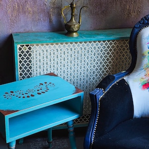 Può includere: Un tavolino laterale blu turchese con un design floreale a stencil si trova accanto a un radiatore blu turchese con un motivo a reticolo bianco. Uno schienale in velluto nero con un cuscino bianco che presenta un ritratto colorato di Frida Kahlo si trova sullo sfondo.
