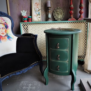 Puede incluir: Una mesa auxiliar redonda verde con tres cajones y patas ornamentadas. La mesa tiene un acabado desgastado y tiradores de cajones de latón de estilo antiguo.