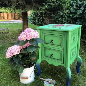 May include: A green painted wooden nightstand with two drawers and decorative floral details on the top. The nightstand has a distressed finish and blue painted legs. The nightstand is sitting on a green lawn with a potted plant and a can of paint.