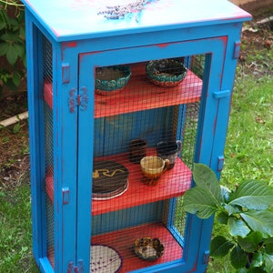 May include: A blue wooden cabinet with a wire mesh front and red shelves. The cabinet has a distressed paint finish and is decorated with small ceramic bowls and cups.