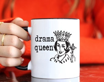 Op de afbeelding: Witte keramische mok met een zwarte rand en handvat. De mok heeft een zwart-witte illustratie van een vrouw met een kroon en een scepter. De tekst "drama queen" is op de mok gedrukt.