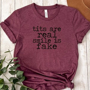 May include: A maroon t-shirt with the text "tits are real smile is fake" printed in black.