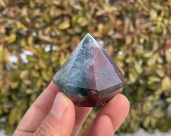 May include: A green, red, and white agate crystal point, also known as a gemstone, in the shape of a diamond. The crystal is being held in a hand.