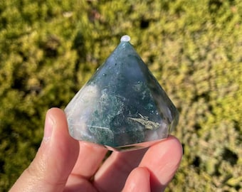 May include: Green moss agate crystal point with a faceted, diamond-shaped design. The crystal is a light green color with white and brown inclusions.
