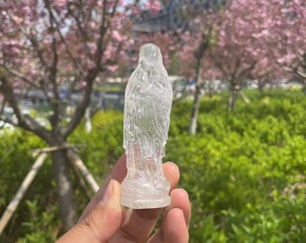May include: A clear crystal statue of the Virgin Mary, carved in a detailed design. The statue is held in a hand against a blurred background of pink flowers and green foliage.