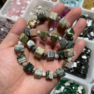 Op de afbeelding: Een armband met vierkant geslepen edelstenen in groen, bruin en crèmekleur. De armband is geregen met kleinere, ronde kralen. De edelstenen hebben een natuurlijk uiterlijk. De armband wordt in een hand gehouden.