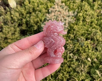 May include: A carved pink seahorse figurine. The seahorse is detailed with a textured body, a curved snout, and a flowing mane. The figurine is held in a hand against a backdrop of green foliage.