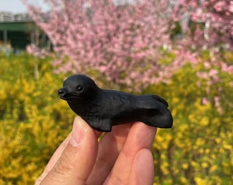 May include: A black carved seal figurine, held in a hand, against a blurred background of pink and yellow flowers.