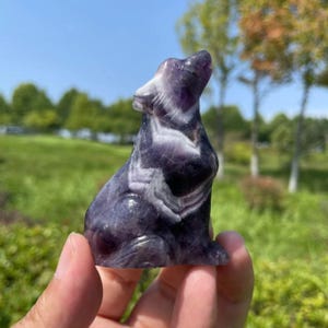 Può includere: Una statuetta di lupo in ametista scolpita con un motivo a spirale viola e bianco. Il lupo è seduto con la testa inclinata all'indietro e la bocca aperta.