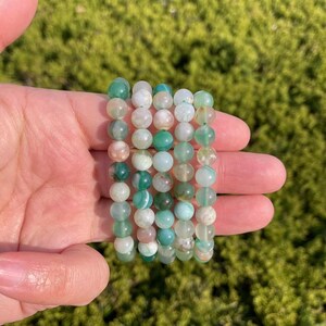 Puede incluir: Una pila de pulseras de ágata verde con acentos blancos y rosados. Las pulseras están hechas de cuentas redondas y están apiladas unas sobre otras.